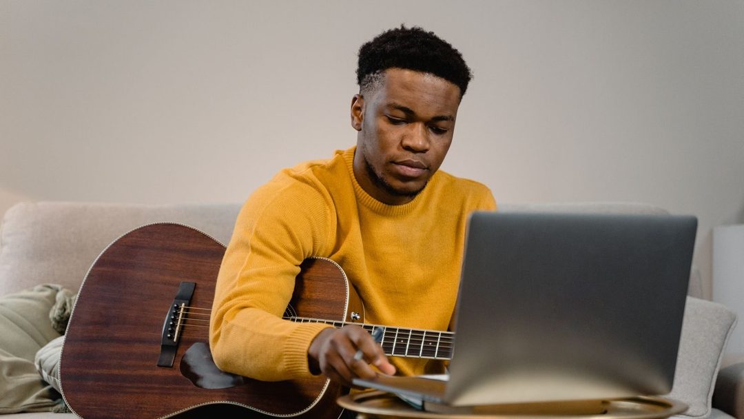 Homem de suéter amarelo toca violão em casa acompanhando uma aula no notebook