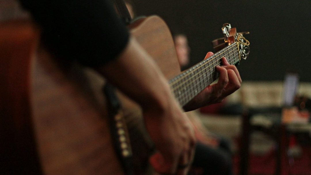 Mão formando um acorde no braço do violão em um ambiente descontraído de café.