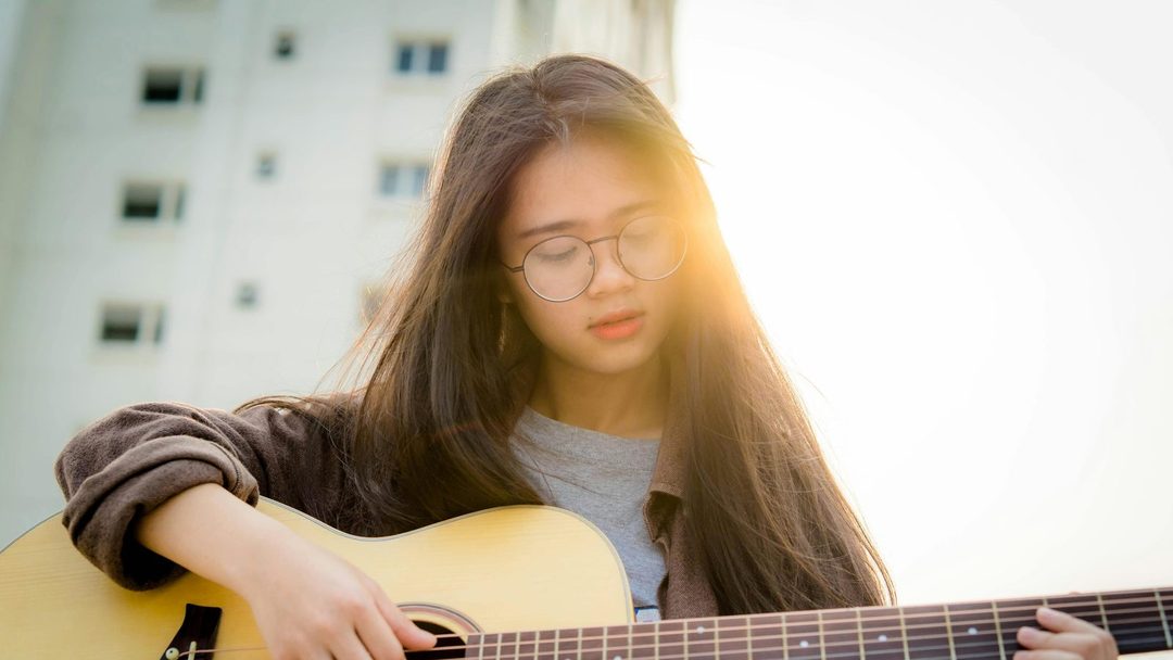 Jovem tocando violão perto da janela, com luz suave iluminando a cena.