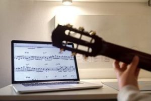 Aluno pratica violão acompanhando uma aula exibida no notebook